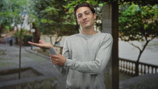 Man presenting palm up with open hand and wrist gesture, wearing glasses and grey sweater on a street; welcoming ease.