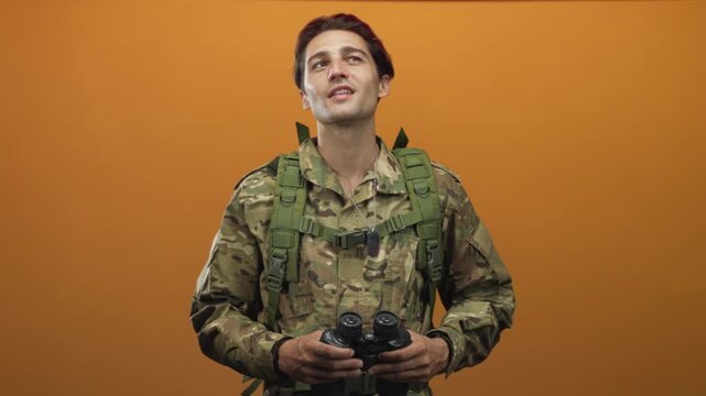 Soldier man in camouflage uniform holding binoculars and dog tags, grip on optics, face turned upward and looking into distance in orange studio; contemplative duty.