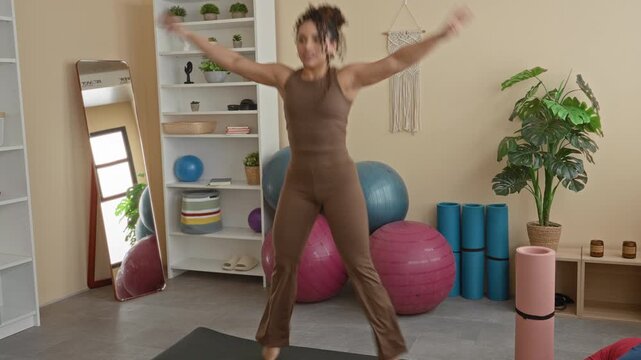 Woman doing jumping jacks with arms raised on yoga mat near exercise ball and foam roller in studio; wellness energetic.