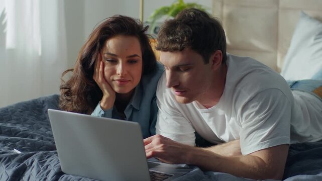Happy Caucasian couple man woman using laptop on bed at home bedroom talking computer online boyfriend girlfriend watching movie female male smiling laugh romance relax love device chatting sweetheart
