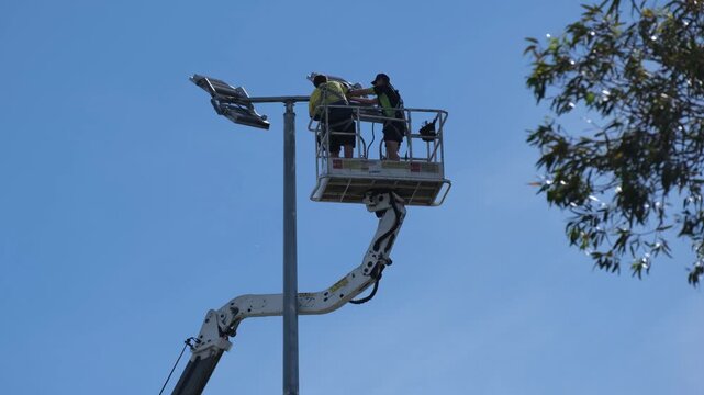Workers using an elevated boom lift to install or maintain sports field lighting in Australia. Infrastructure maintenance scene highlighting electrical work, safety procedures, public facility upkeep