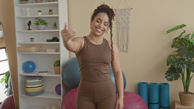 Woman smiling extends her hand toward the camera beside large exercise balls and foam rollers in a bright studio with plants, a shelf and macrame wall hanging; welcoming calm.