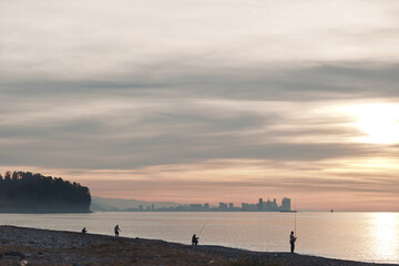 Obraz premium Beach sunset with silhouettes of fishermen along the shore, calm water and distant skyline, warm glow and golden light creating a tranquil coastal scene for travel and nature photography.