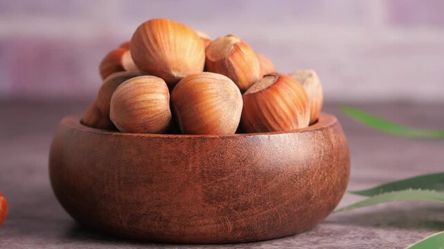 Close up of whole hazelnuts in a brown bowl with green leaves on a dark table.