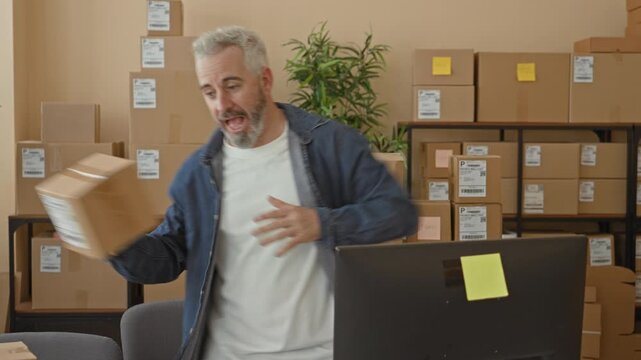 Man throws parcel with hand over computer monitor in building near stacked boxes and plant; frustration small business logistics.