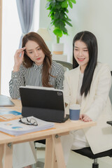 Two Asian businesswomen sat opposite each other at a desk, each using a laptop or tablet to analyze data. On the desk were neatly organized documents with charts and graphs related to the analysis.