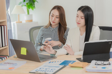 Two Asian businesswomen sat opposite each other at a desk, each using a laptop or tablet to analyze data. On the desk were neatly organized documents with charts and graphs related to the analysis.