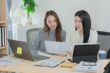 Two Asian businesswomen sat opposite each other at a desk, each using a laptop or tablet to analyze data. On the desk were neatly organized documents with charts and graphs related to the analysis.