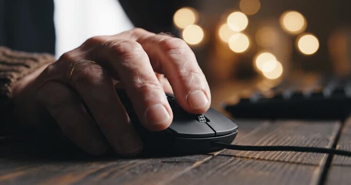 Close-up of an elderly person's hand using a computer mouse on a wooden desk with soft bokeh lights