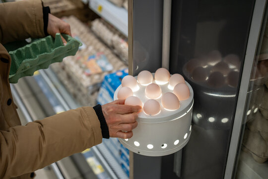 Unrecognizable woman in supermarket putting raw egg from box inside special ovoscope to check quality of eggs by candling light through shell. Person examinees condition of eggs in grocery store.