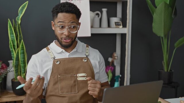 Man florist in brown apron writes order on paper beside open laptop with tulips and vases on shelf in studio; focused small business.