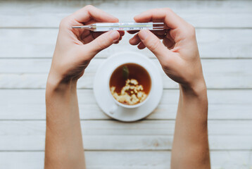 Normal temperature. Woman's hands holding a mercury thermometer on a white wooden background.Concept of colds, flu, and acute respiratory infections.Linden blossom tea as an alternative cold treatment