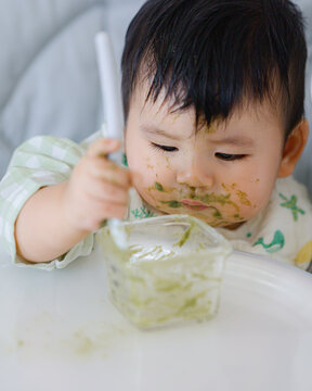 Happy Asian baby boy in high chair smiling at mother while being spoon-fed. Close-up view with natural light in a home setting.	
