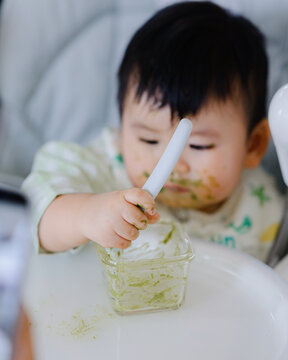 Happy Asian baby boy in high chair smiling at mother while being spoon-fed. Close-up view with natural light in a home setting.	
