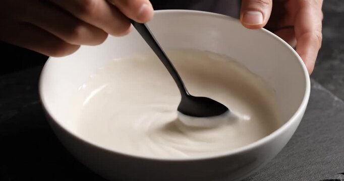 Close-up of hands stirring creamy mixture in a white bowl on a dark surface, with soft lighting
