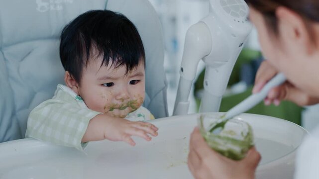Happy Asian baby boy in high chair smiling at mother while being spoon-fed. Close-up view with natural light in a home setting.	

