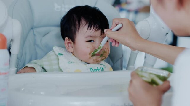 Happy Asian baby boy in high chair smiling at mother while being spoon-fed. Close-up view with natural light in a home setting.	

