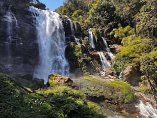 Obraz premium Majestic Wachirathan Waterfall cascades powerfully down a multi-tiered rock face, surrounded by vibrant tropical foliage and sunlit mist in Doi Inthanon National Park, Chiang Mai, Thailand. A stunning
