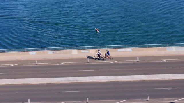 Two cyclists in helmets ride bikes along scenic waterfront path at Mission Bay Park San Diego while kayaker paddles in calm turquoise water.