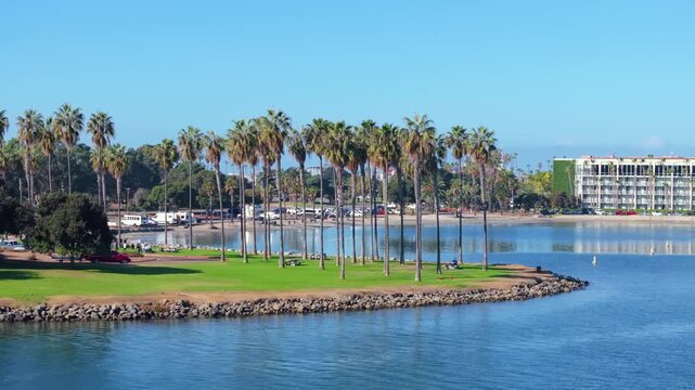 Scenic aerial view of Mission Bay Park island in San Diego California featuring tall palm trees on lush green grass peninsula surrounded by calm blue water with resort buildings and beach in backgroun
