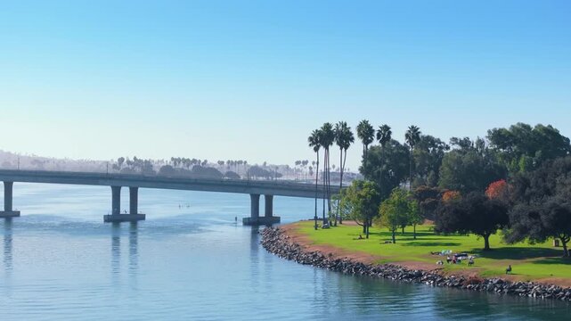 Aerial panoramic view of Mission Bay Park in San Diego, California. Concrete bridge spans calm blue water with paddle boarders below. Lush green park with tall palm trees and rocky shoreline.