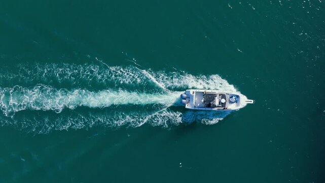 Drone footage captures white motorboat speeding across deep turquoise sea water, leaving foamy wake trail behind. Boat passes under bridge with cars driving on highway.