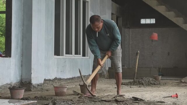 Front view of a construction worker leveling the ground using a traditional blencong pickaxe while smoking