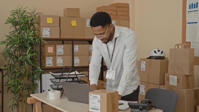 Man leans on desk over laptop and parcel amid stacked boxes and shelf in building mailroom; focused productivity.