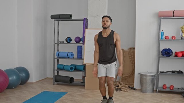 Man in black tank top and white shorts raises arms overhead performing a jumping jack on a blue mat in a fitness studio with exercise balls and ropes visible; energy fitness focus.