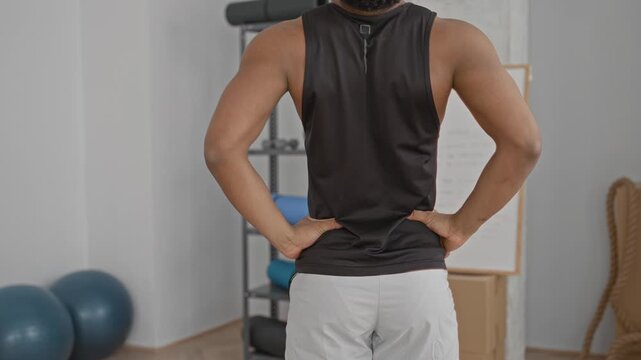 Man with bare back and muscular arms, hands on hips standing facing gym equipment with exercise balls and foam rollers in a training studio; determination focus resilience.