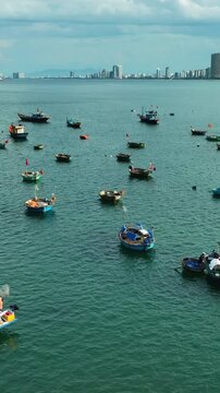 Vertical video,Colorful fishing boats anchored on calm sea at Vietnam beach with coastal city skyline, traditional round basket boats and peaceful seaside atmosphere under soft daylight.
