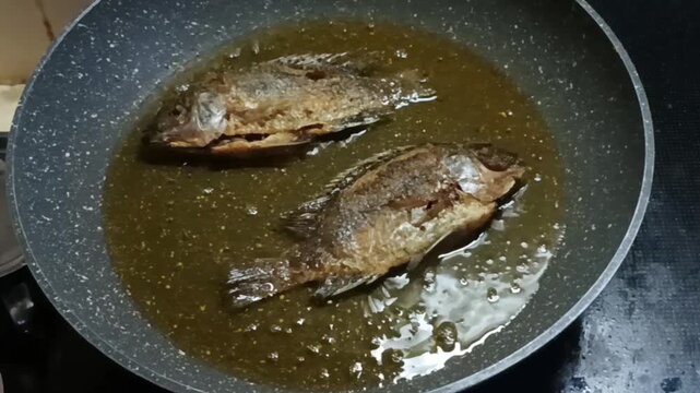 Whole tilapia fish are being fried in a pan filled with hot oil on the stove