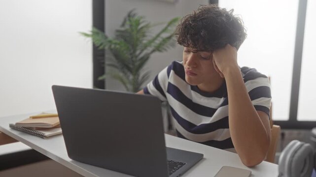 Student boy in striped shirt covers mouth yawning at open laptop on white desk inside a building with notebooks stacked nearby; late night fatigue.