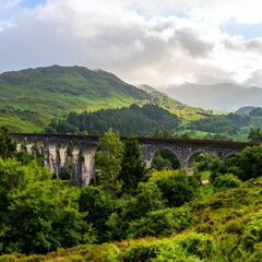 Fototapeta premium Glenfinnan Viaduct in Scottish Highlands, iconic railway bridge amidst lush green landscape and dramatic cloudy sky.
