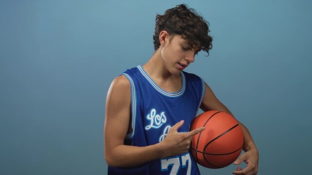 Teen boy in blue los angeles jersey holding basketball and beckoning with hand in studio; confidence swagger.