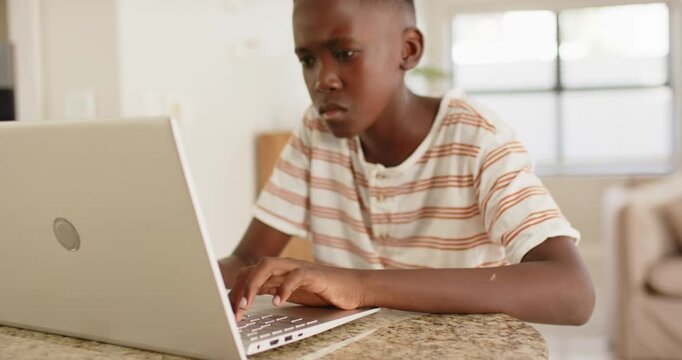 Teenage male in striped shirt typing on silver laptop at granite counter, pausing doing homework