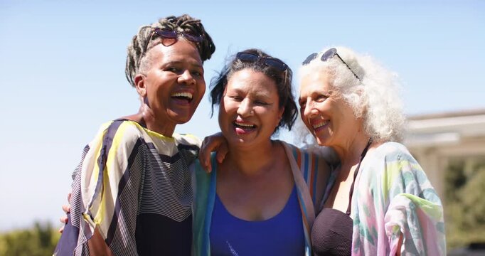 Diverse female friends camera cue posing at park laughing, shifting, wearing shades and swimsuits