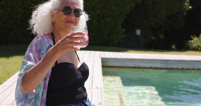 Camera panning showing woman in swimsuit shorts on pool deck holding pink drink sipping relaxing