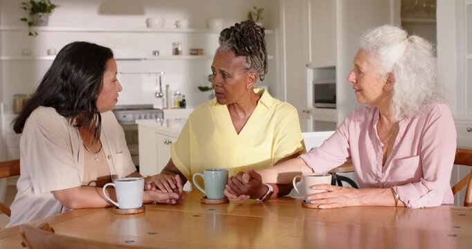 Three female friends at dining table with mugs, left woman speaking, holding hands for support