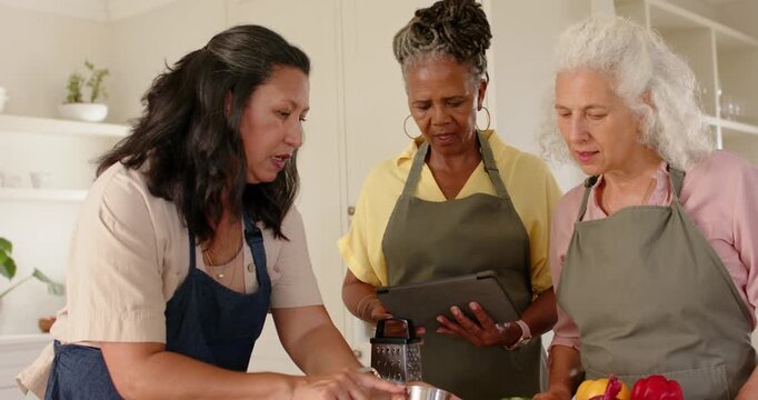 Diverse female friends leaning arranging prep and measuring cups at kitchen island checking tablet