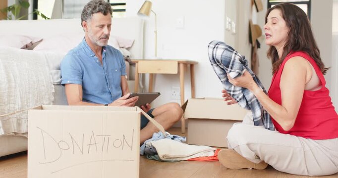Couple checking tablet list for donation, folding and packing shirts into donation box in bedroom