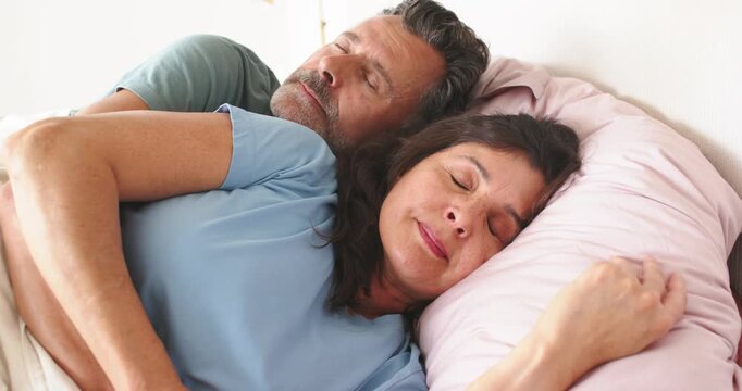 Mature couple lying on bed, man cradling woman while morning light waking her, pale pink pillow