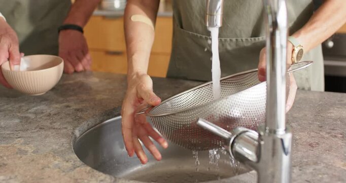 Diverse couple; woman in apron rinsing tomatoes in colander with faucet running, man holding bowl