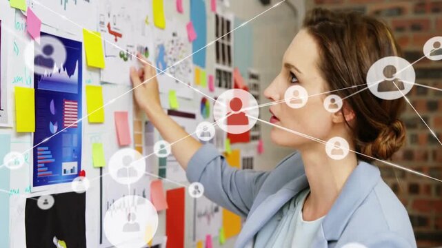 Female marketing planner moving sticky note at office, causing network icons expanding over graphs