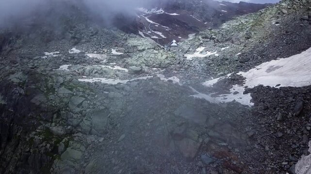 Drone flies toward Sach Pass, a remote Himalayan mountain pass in Himachal Pradesh, revealing rocky slopes, snow patches, low clouds, and a 4x4 vehicle at the summit.