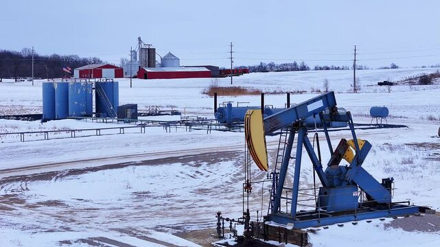 Oil pumpjack operating near farm buildings in snowy Michigan, USA field