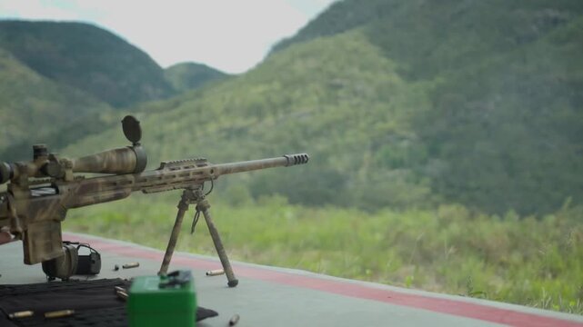 A rifle set up on top of a mountain range fires with a puff of gun powder and smoke coming out of the barrel.