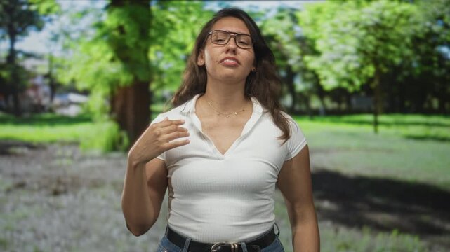 Woman pinches nose in a sunny green park among trees and grass, wearing glasses and a white shirt; disgust avoidance.