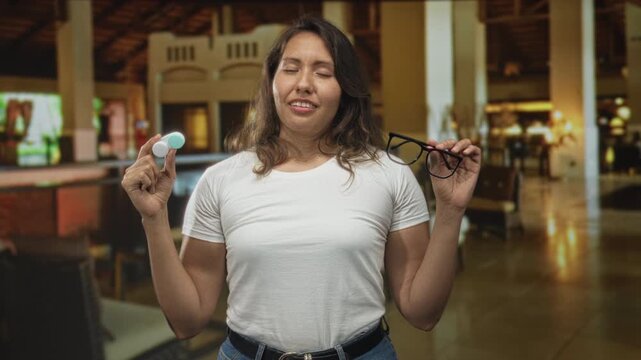 Woman holding glasses and contact lens case in hotel building lobby, eyes closed and smiling with hands raised; relief travel.