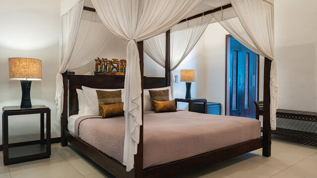 A large bed with a canopy and pillows is in a room with a white wall. The bed is surrounded by a white curtain, and there are two lamps on either side of the bed